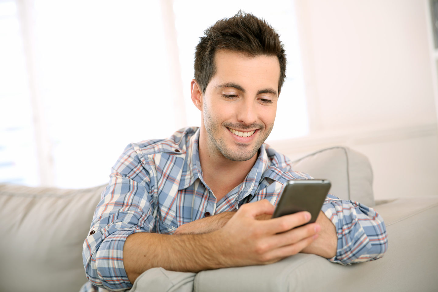 Un homme regarde son téléphone en souriant, sur son canapé